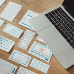 Een laptop op een houten bureau naast verschillende handgetekende draadframeschetsen van websites in blauwe en rode inkt, ter illustratie van de stap voor stap proces van website bouwen en website lanceren.