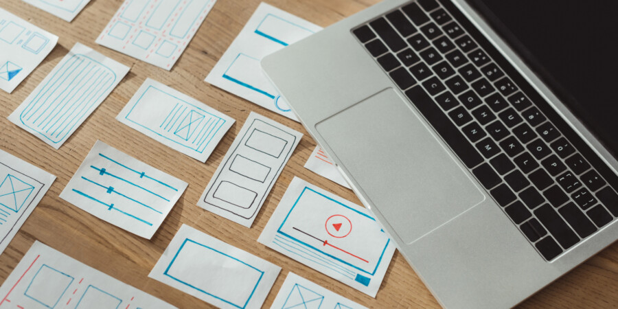 Een laptop op een houten bureau naast verschillende handgetekende draadframeschetsen van websites in blauwe en rode inkt, ter illustratie van de stap voor stap proces van website bouwen en website lanceren.